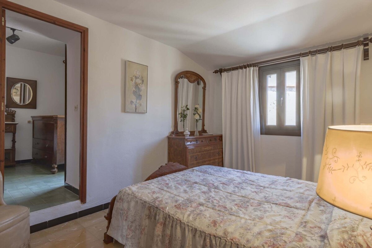 A bedroom with a floral bedspread, wooden dresser with an ornate mirror, a window with white curtains, and an open doorway to another room.