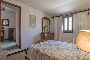 A bedroom with a floral bedspread, wooden dresser with an ornate mirror, a window with white curtains, and an open doorway to another room.
