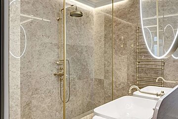 Luxurious bathroom with gray marble walls, brass shower fixtures, and double sinks. Features a glass partition, heated towel rack, and illuminated mirror.