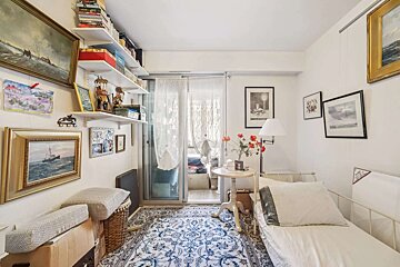 A cozy, eclectic room featuring a single bed, ornate rug, shelves with books and decor, many framed pictures, and sheer curtains leading to a balcony.