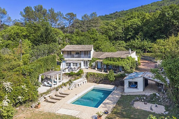 An aerial view of a hillside villa with a swimming pool, sun loungers, and lush green trees under a clear blue sky.