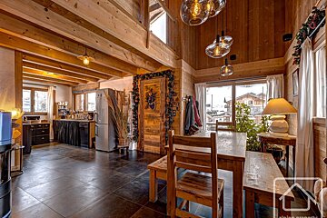 Cozy wooden chalet interior featuring a dining area, exposed beam ceiling, decorative hanging lights, and a kitchen in the background.