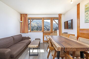 Bright living/dining room with a brown sofa, rustic wooden furniture, and large windows showcasing a stunning mountain village view.