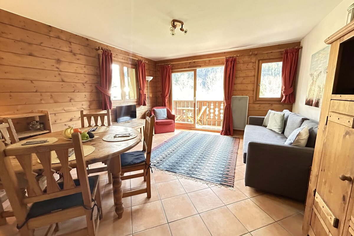 A cozy, wood-paneled living room features a dining table, sofa, red armchair, and a balcony, all on a tiled floor with a patterned rug.