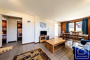 Cozy ski chalet-style living/dining area with tiled floors, wood-paneled walls, a blue sofa, dining table, and TV. Doorways lead to other rooms.