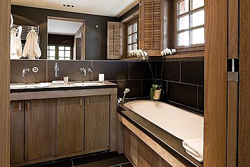 Modern bathroom with dark tiled walls, wooden double vanity, white bathtub, and orchids by shuttered windows.