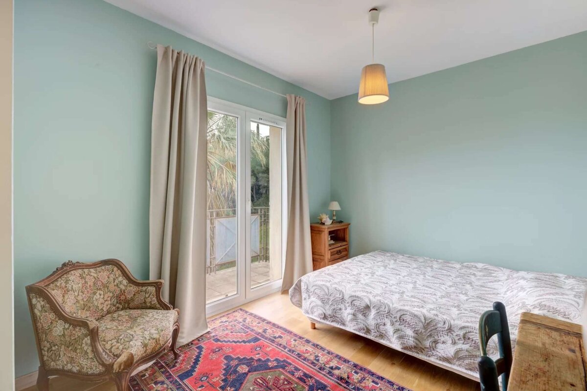 A cozy bedroom with light blue walls, a bed, floral armchair, red patterned rug, and French doors leading to a balcony.