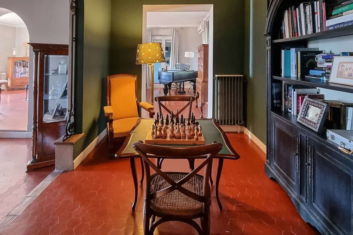 A classic room with dark green walls, terracotta floor, and a chessboard on a table. A large bookshelf, a cozy chair, and a peek into a piano room complete the scene.