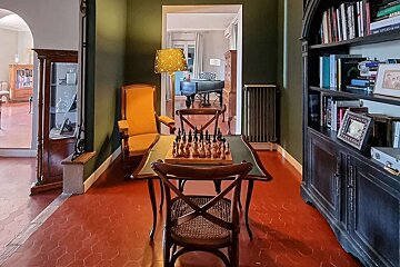 A classic room with dark green walls, terracotta floor, and a chessboard on a table. A large bookshelf, a cozy chair, and a peek into a piano room complete the scene.