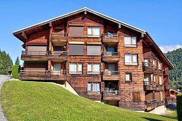 A large multi-story wooden chalet with balconies and many windows sits on a green hill against a clear blue sky and mountains.