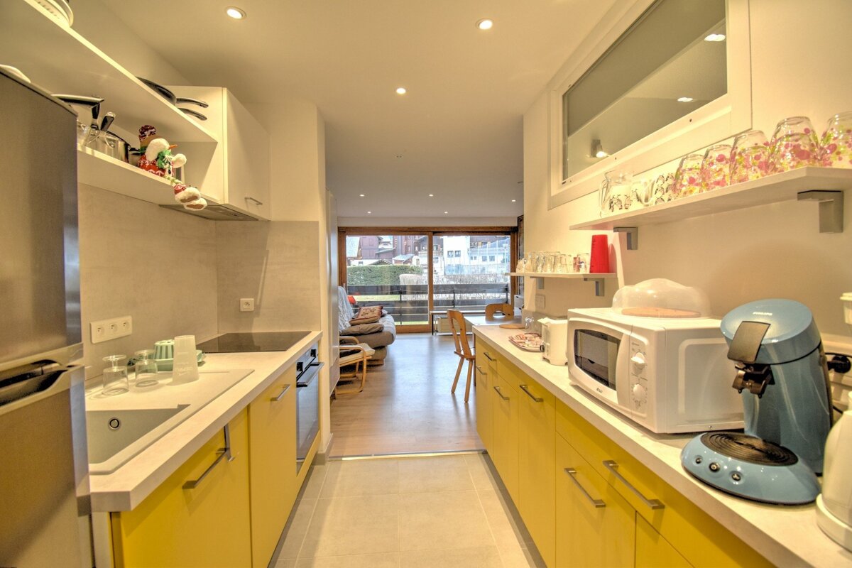 A modern, narrow galley kitchen featuring bright yellow cabinets, white counters, and appliances, leading to a living area with a balcony view.