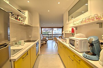 A modern, narrow kitchen with bright yellow cabinets, white countertops, and appliances. It opens up to a living area with a balcony view.