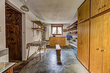 A rustic storage room or pantry with a concrete floor, white walls, wooden cabinets, and shelves. A table, bench, and window are also visible.