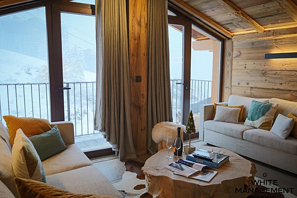 A cozy, luxurious ski chalet living room with plush sofas, a unique wood coffee table, and a snowy mountain view. Champagne awaits in this rustic space.