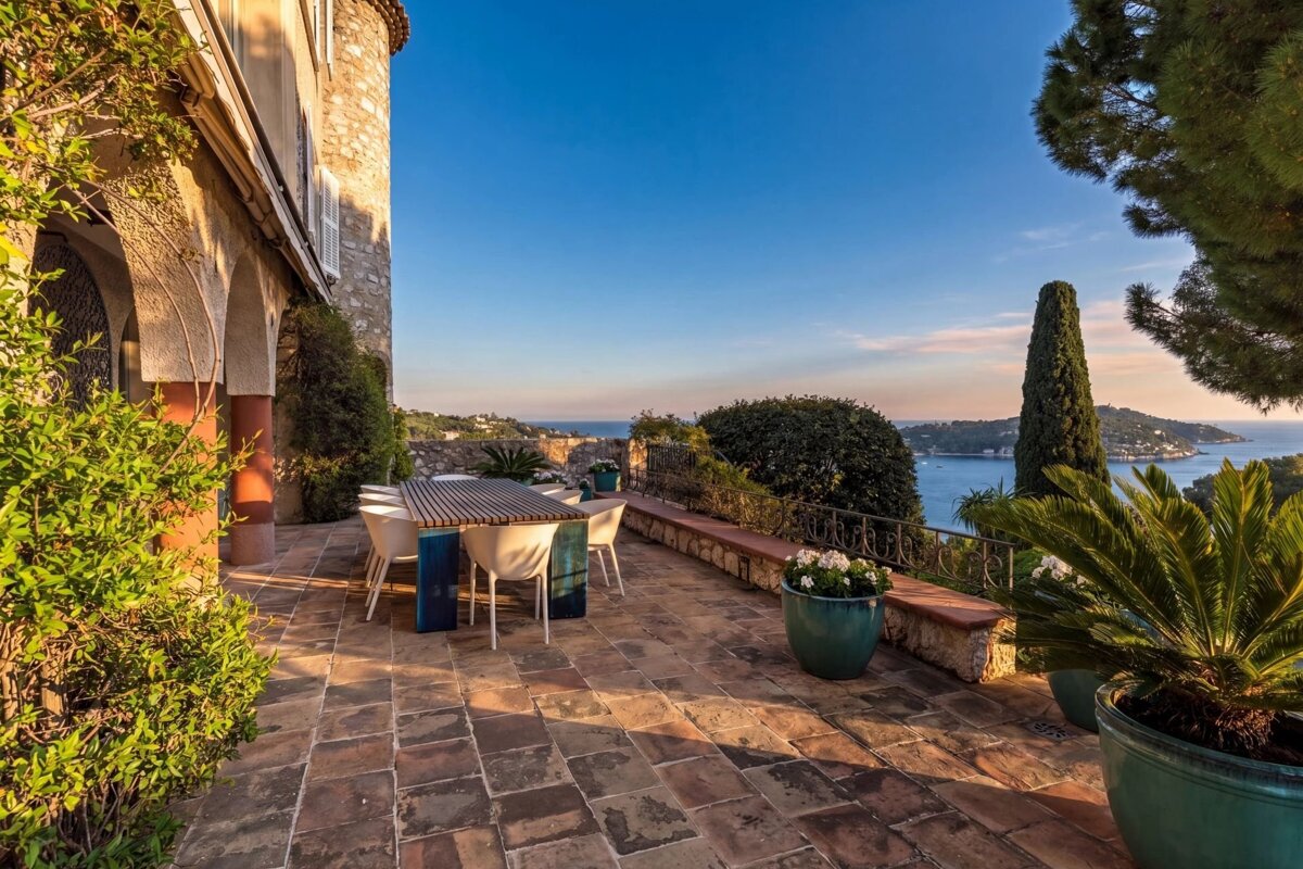 A picturesque stone terrace with an outdoor dining table, bathed in warm sunlight, offers panoramic views of the tranquil Mediterranean sea and coastline.