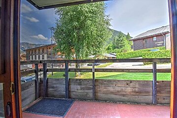 View from a wooden balcony showing a green lawn, chalet-style buildings, trees, and mountains in the distance.