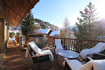 A sunny wooden deck with a hot tub and cozy seating, overlooking a snowy mountain village. Ideal for winter relaxation.