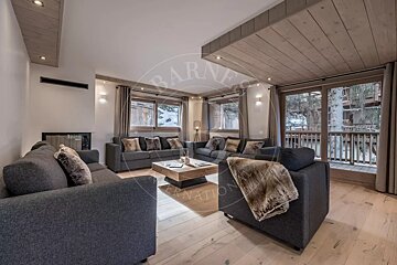 A chic, modern living room featuring grey sofas with fur accents, light wood floors/ceiling, and a fireplace. Large windows offer views of a snowy exterior with trees.