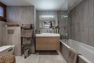 A modern bathroom with grey wood-look tiles, a floating wood vanity, bathtub with shower screen, toilet, and towel warmer, featuring neutral tones.