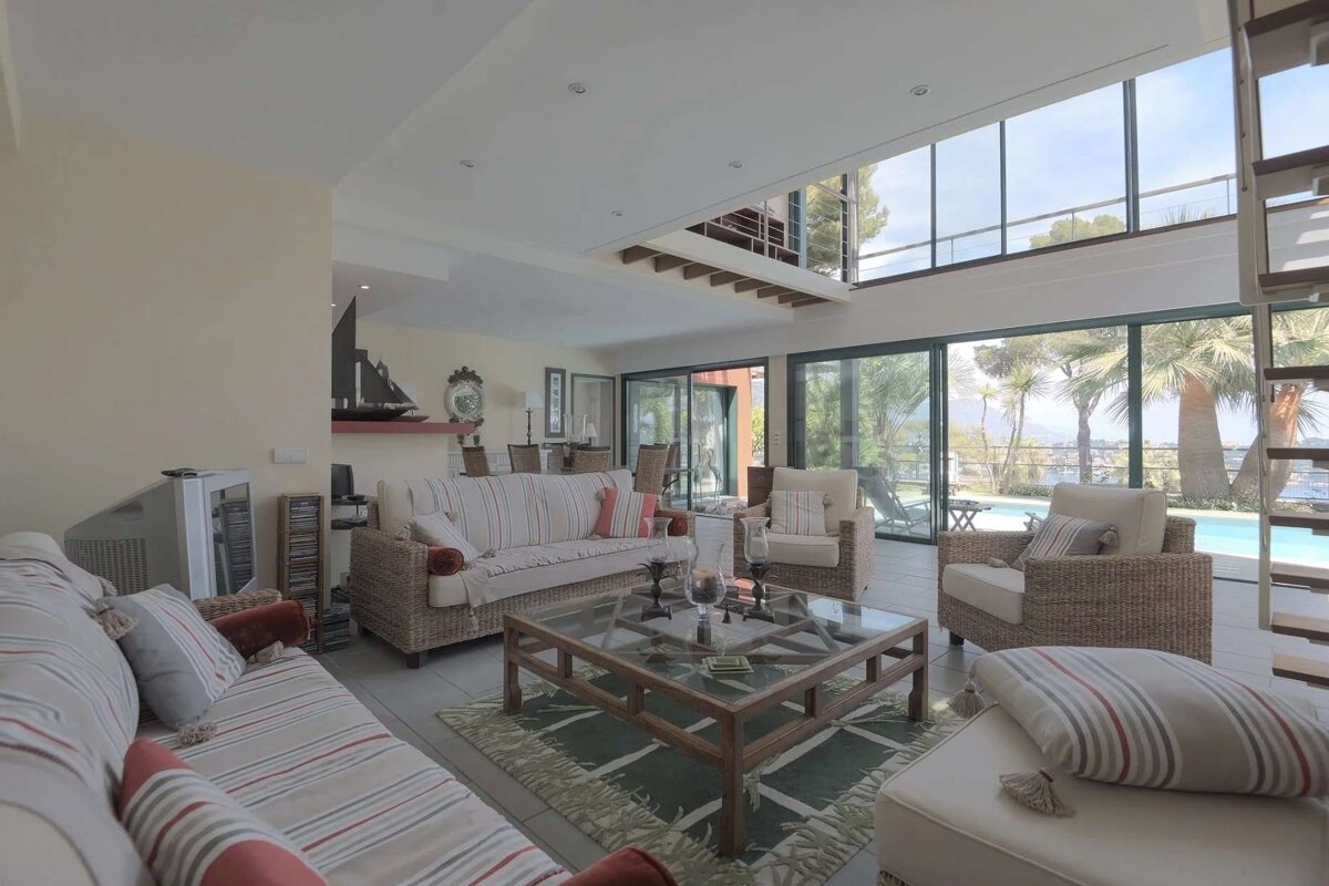 A bright, spacious living room with large windows offering a pool and sea view. Features coastal-style furniture, high ceilings, and a modern design.