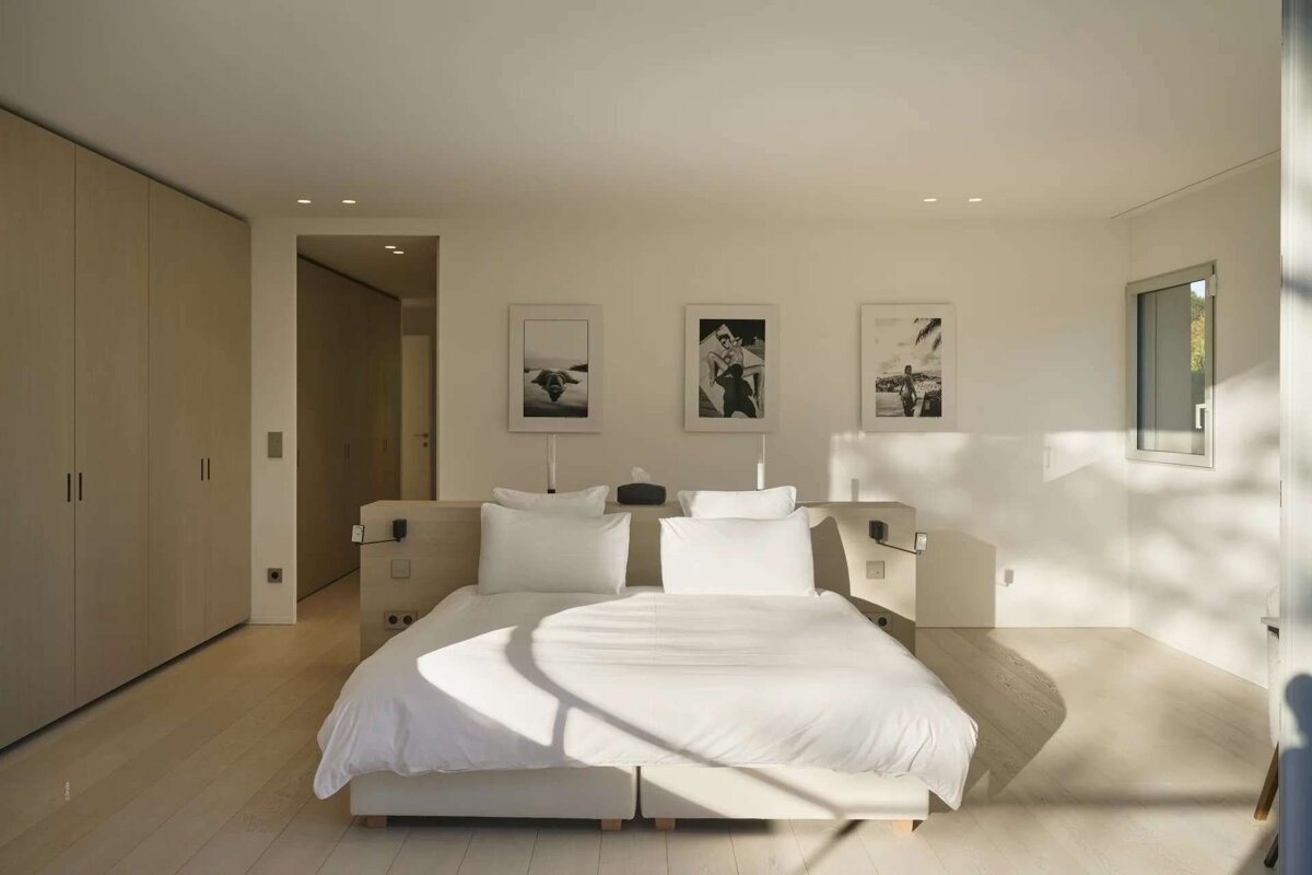 Bright, minimalist bedroom with a white bed, light wood floors and wardrobes. Sunlight creates geometric shadows, enhancing the serene, modern aesthetic.