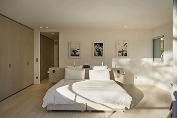 Bright, minimalist bedroom with a white bed, light wood floors and wardrobes. Sunlight creates geometric shadows, enhancing the serene, modern aesthetic.