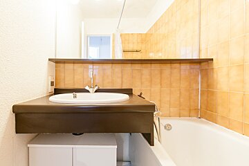A bathroom featuring a dark brown sink counter with a white basin, a large mirror, light brown tiled wall, and a white bathtub. A small cabinet sits below.