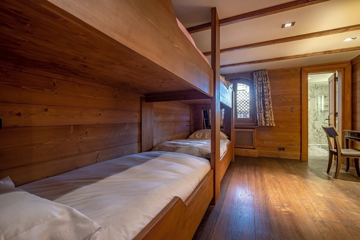 A cozy, wood-paneled room with wooden bunk beds, a window with patterned curtains, and an open doorway leading to a marble-tiled bathroom.