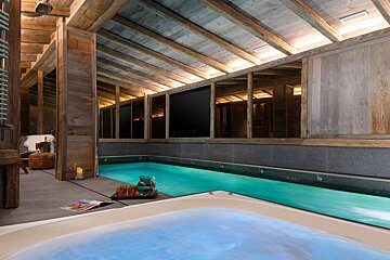 A luxurious, rustic indoor pool and hot tub in a wooden chalet, featuring warm lighting, a cozy ambiance, and refreshments nearby.