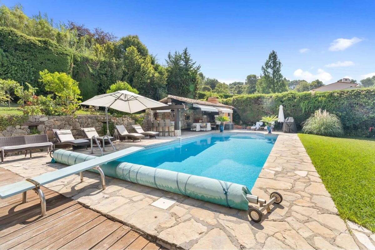 A beautiful backyard pool with a diving board, sun loungers, an outdoor kitchen, and vibrant green landscaping under a sunny blue sky.
