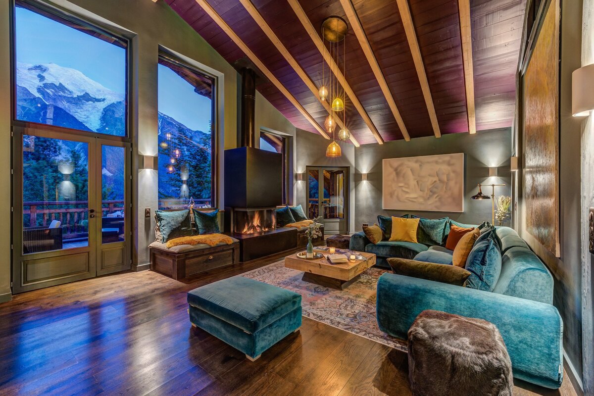 Luxurious, cozy living room with panoramic snowy mountain views. Features a modern fireplace, teal velvet seating, dark wood floors, and a beamed ceiling.