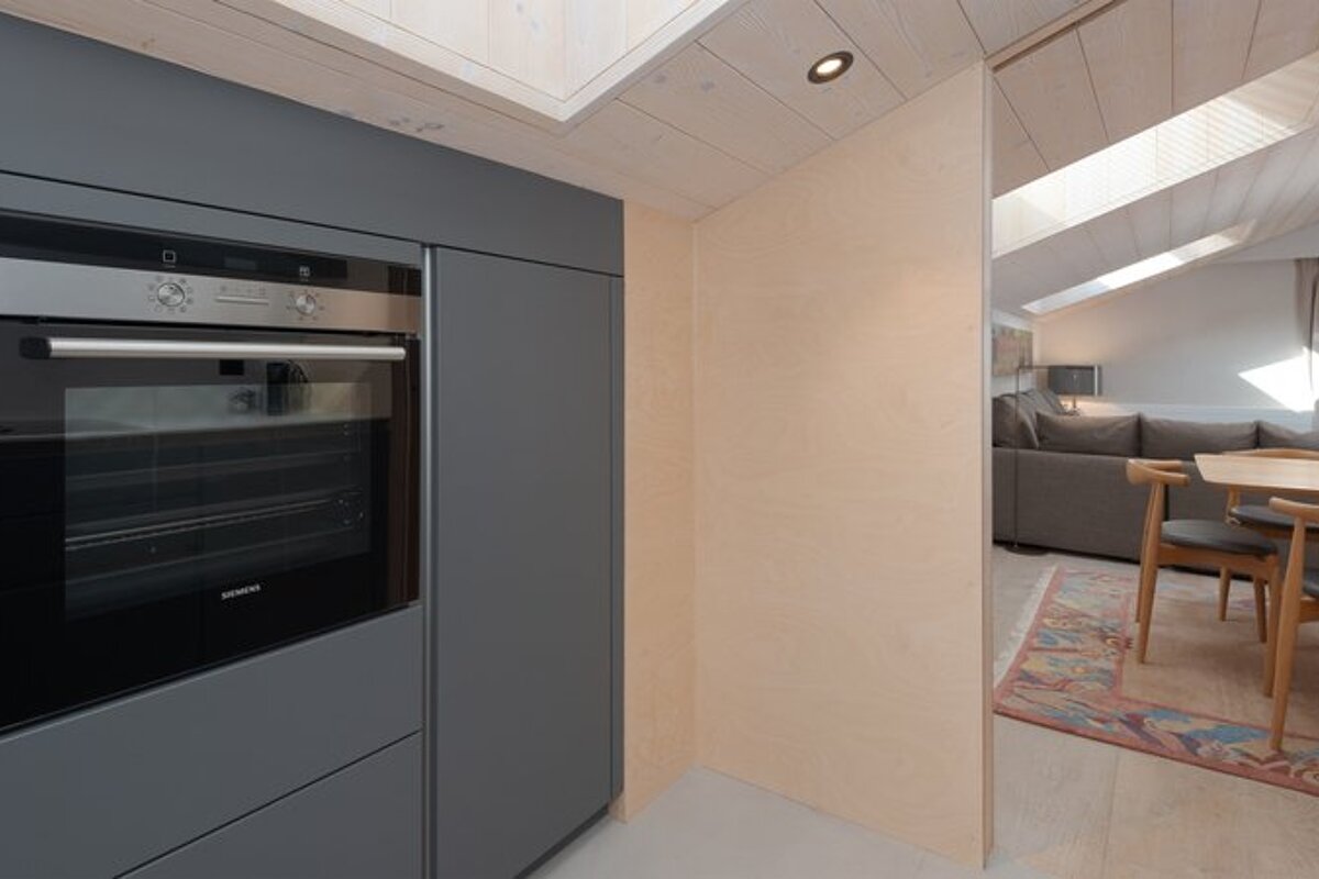 Modern gray kitchen cabinets with a built-in oven next to light wood walls, looking into a cozy living room under a slanted roof.