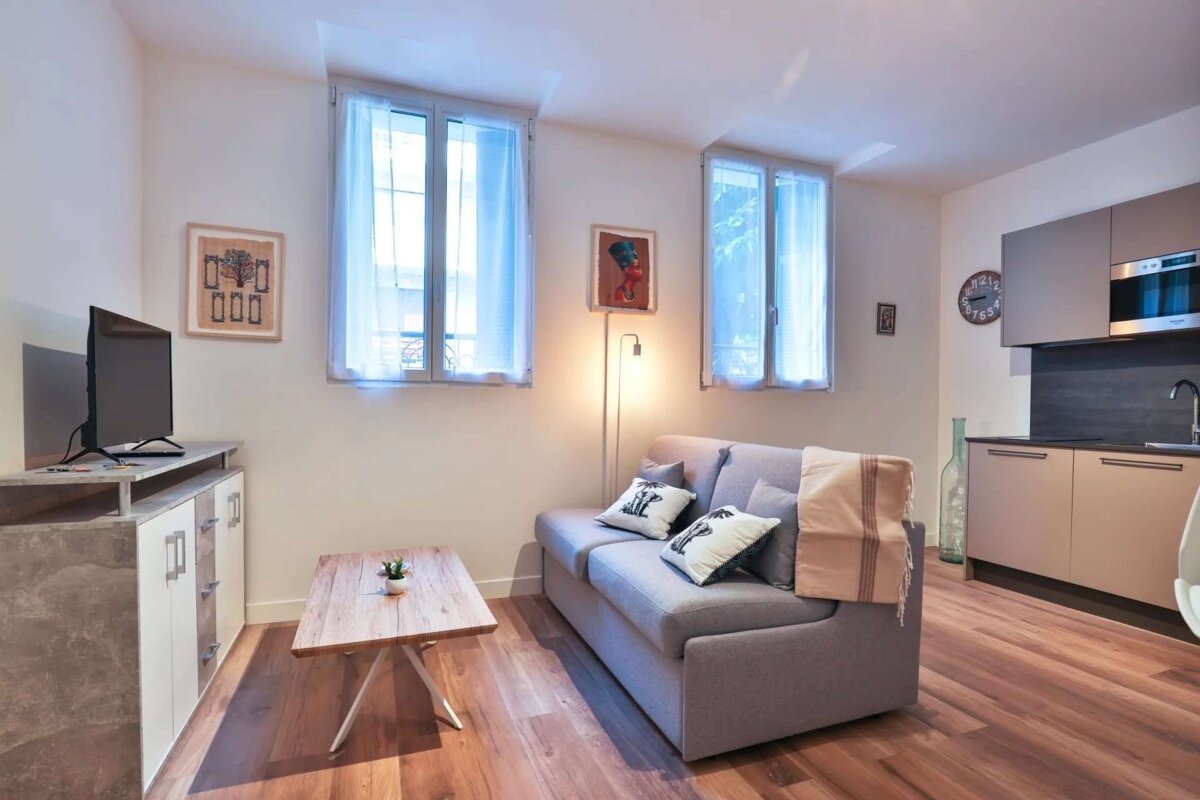 A bright, modern studio apartment with a gray sofa, TV, wooden floors, and a small kitchenette. It features large windows and subtle decor.