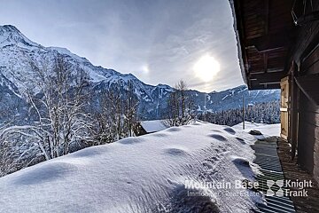 A bright winter scene with snow-covered mountains, frosted trees, and a sun-drenched valley, viewed from a snowy slope beside a rustic wooden building.