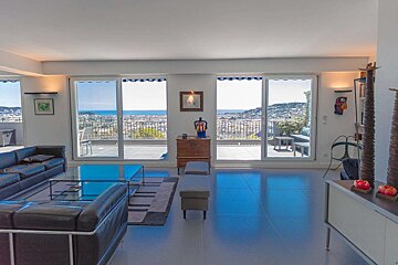 Spacious living room with large glass doors offering panoramic sea and city views from a terrace. Features black leather sofas and modern decor.