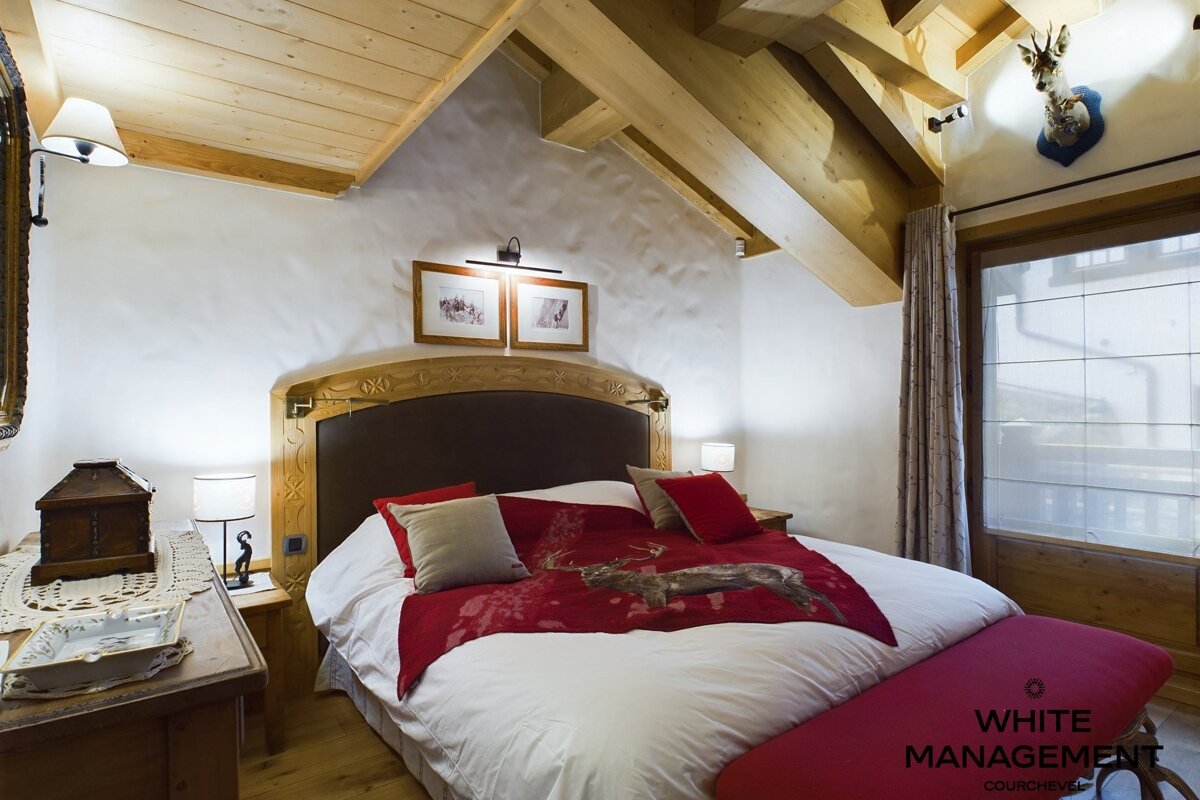 A cozy, rustic chalet bedroom with wooden beams, white walls, and a large bed featuring red bedding with a deer design. A deer head decorates the wall.