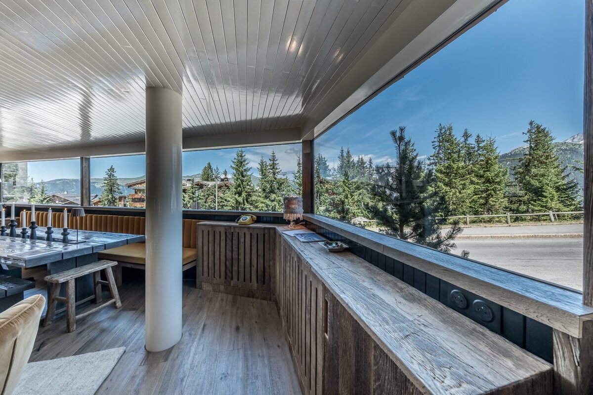 Modern rustic chalet interior with large panoramic windows overlooking a scenic pine forest and mountain road under a blue sky.