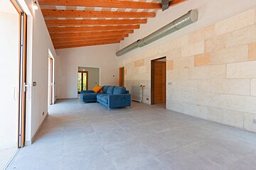 A large, bright room featuring a blue sectional sofa, exposed wooden ceiling beams, natural stone wall, and grey tiled floor, with an industrial vent.