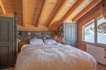 Cozy wooden cabin bedroom with a fluffy white blanket on the bed and a window showing a snowy winter landscape outside.