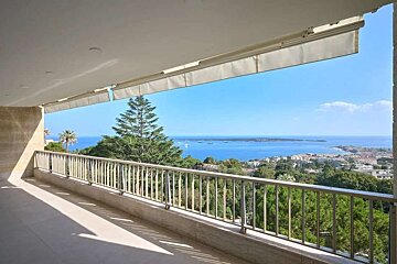 A spacious balcony offers a panoramic view of a coastal town, a vibrant blue sea dotted with boats, and lush green trees under a clear sky.