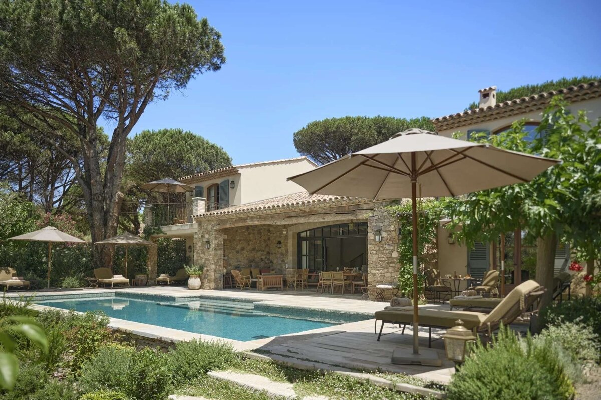 A luxurious stone villa with a swimming pool, lounge chairs, and umbrellas under a bright blue sky, surrounded by lush green trees and plants.