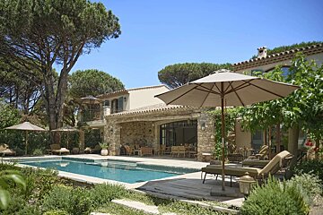 A beautiful stone villa with a sparkling swimming pool, shaded loungers, and lush trees under a bright blue sky, creating a serene outdoor oasis.