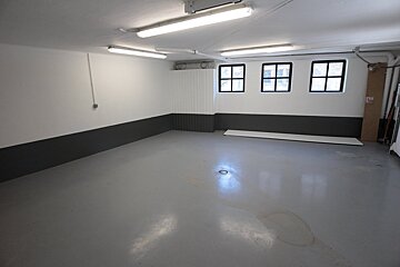 Empty, brightly lit room with white and dark gray walls, gray floor, fluorescent lights, and three small grid windows.