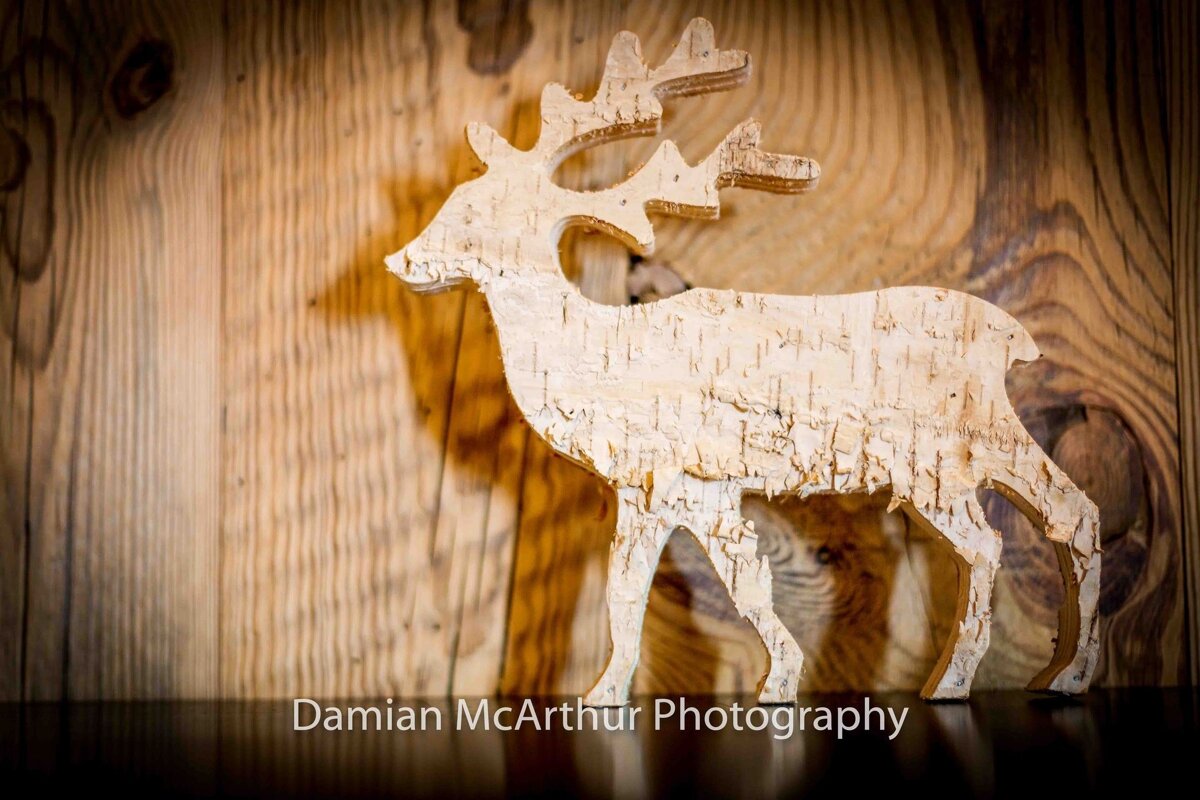 A rustic birch bark cutout of a reindeer stands on a reflective surface against a wooden wall.