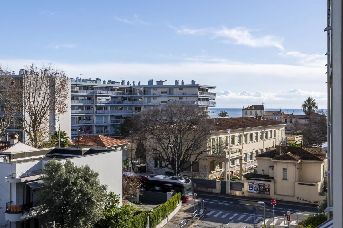 An elevated view of a coastal city, showing multi-story apartment buildings, trees, a street with a bus, and the sea on the horizon.