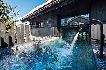 An outdoor spa pool with a waterfall, nestled beside a snowy wooden chalet under a clear sky, offering a luxurious mountain retreat.