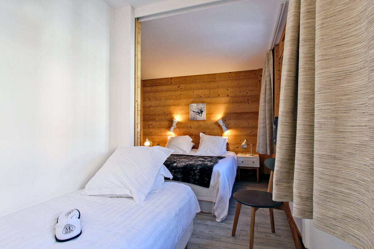 A cozy bedroom featuring a main bed against a wood-paneled wall, a smaller single bed, and light-colored curtains.