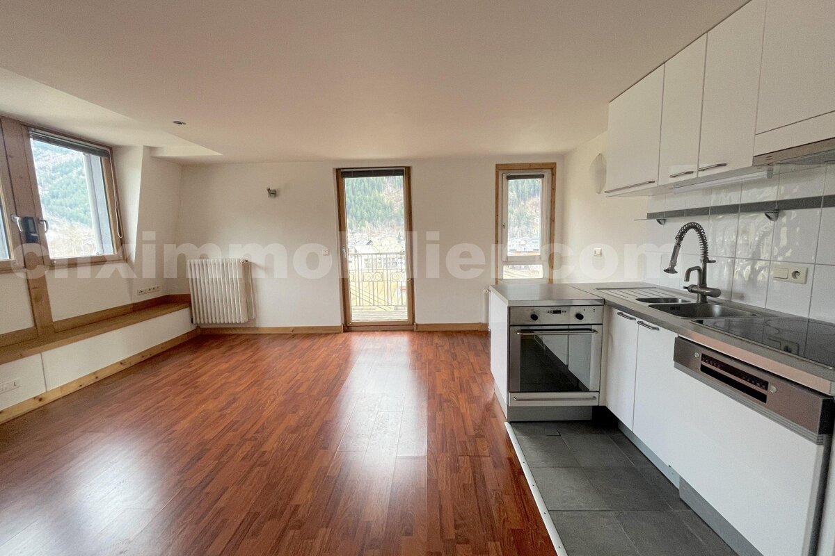 Bright, open-plan living room with wood floor, large windows, and a modern kitchen featuring white cabinets, oven, and sink.