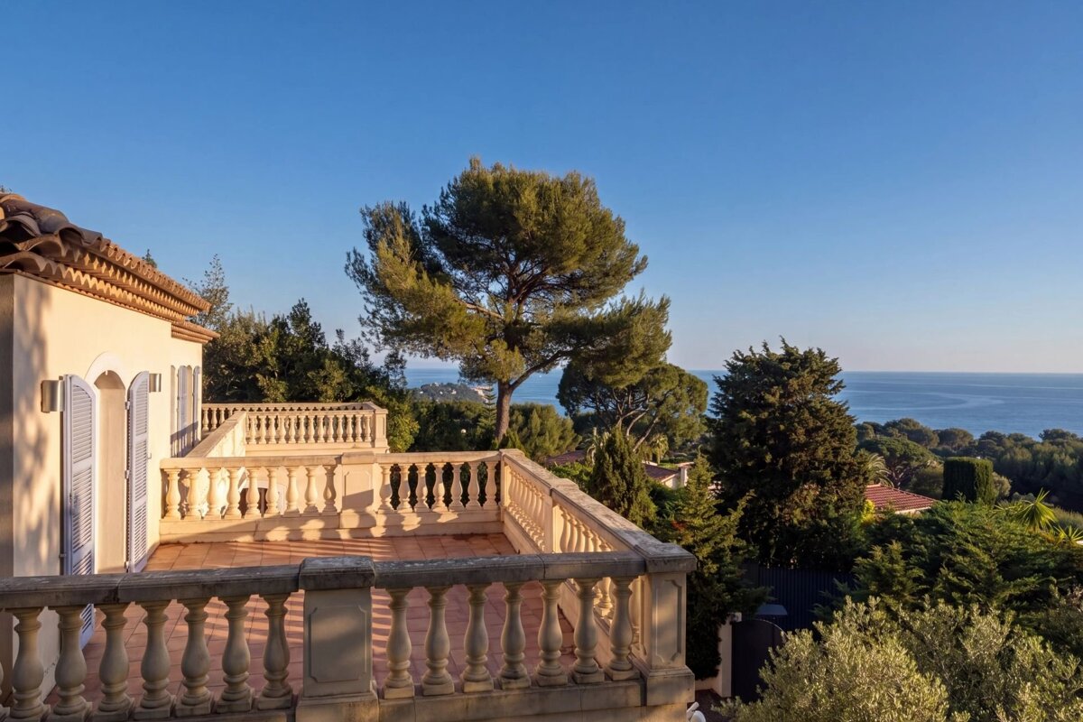 A stone balcony of a villa overlooking green trees and the blue sea under a clear sky.