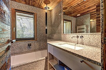 A modern bathroom featuring speckled granite walls & counter, a wooden ceiling, sink with large mirror, and a window with blinds.